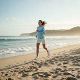 Active woman in light blue and white sportswear running along a sunny beach with ocean waves, representing vitality, movement, and wellness.