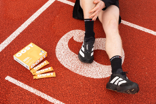 An athlete sitting on a red running track massaging their calf, alongside a box and individual sachets of OOST liquid marine collagen for active recovery.