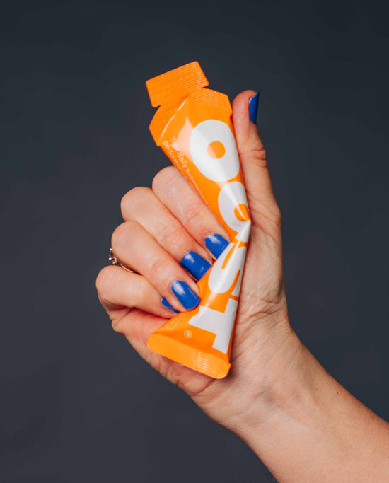 A medium close-up shot of a hand with blue nail polish, holding a single-serving OOST supplement sachet against a dark, solid charcoal background. The white OOST logo is partially visible on the orange packaging, along with the text 'Your daily supplement for performance' and a small 'PEACH' text.